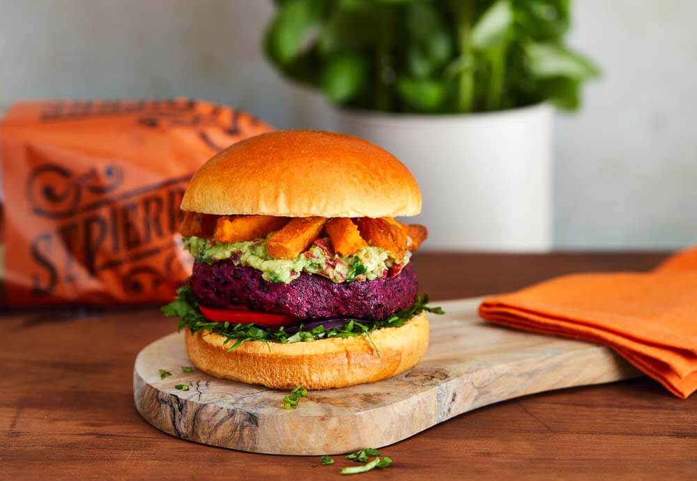 A brioche burger bun filled with a beetroot burger, lettuce, tomato, guacamole and sweet potato fries, on a wooden board