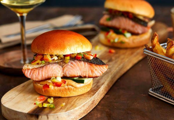 A photo of two burger buns filled with salmon fillets and diced vegetables, on a wooden board