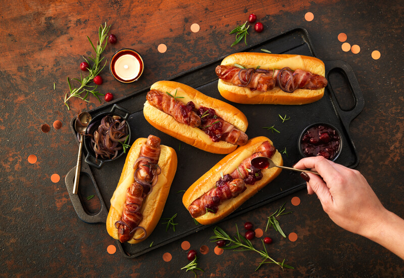 blogpostrecipeinspoxmasbreakfasts An overhead photo of 'Christmas hot dog rolls with a hand adding cranberry sauce on top