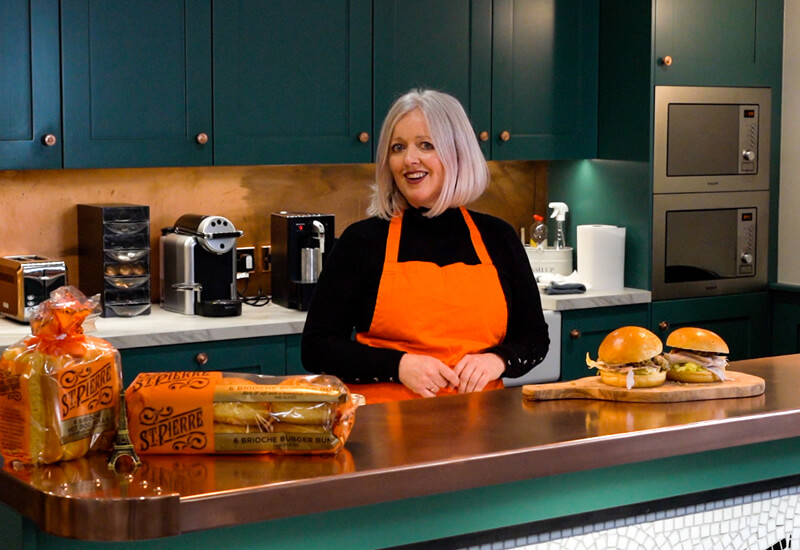 A lady in an orange apron standing in a kitchen behind a kitchen counter with burger buns and hot dog rolls on top