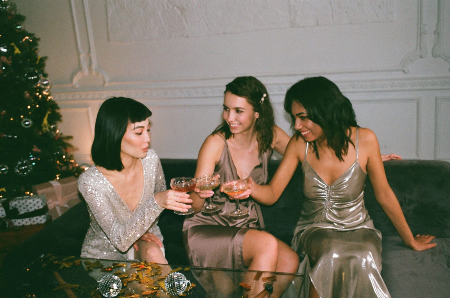Three women sat on a sofa in evening cocktail dresses next to a Christmas tree