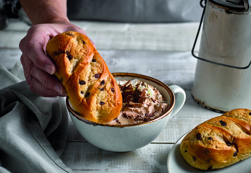 A chocolate chip brioche roll being dunked into a big mug of hot chocolate and whipped cream