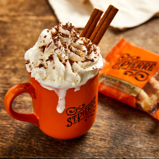 An orange mug with whipped cream and chocolate on top, and two cinnamon sticks sticking out