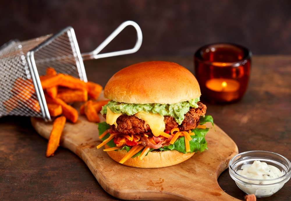 A close up photo of a burger bun filled with fried chicken, salad, melted cheese and guacamole on a wooden board with sweet potato fries