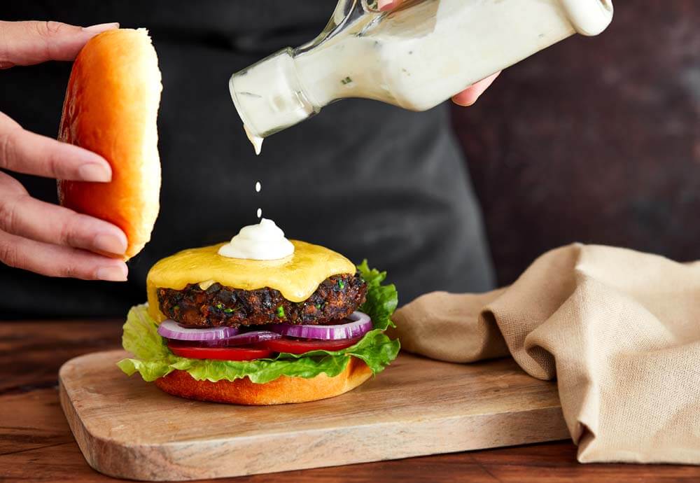 A hand dripping sauce on top of a burger, on a wooden serving board with a tea towel next to it