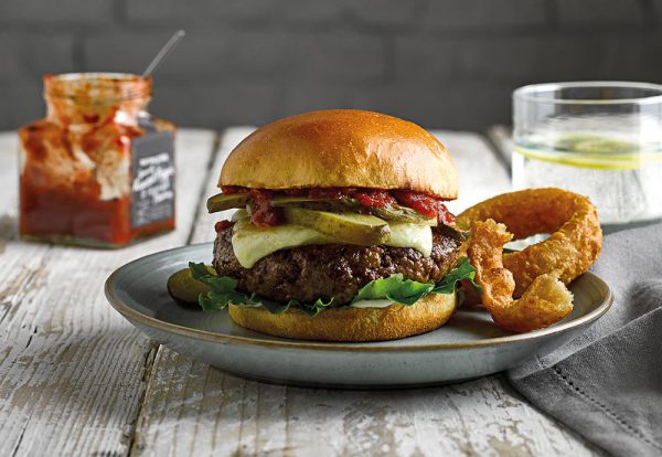 A close-up photo of a beef burger with chutney, pickles and cheese on top, served on a plate with onion rings