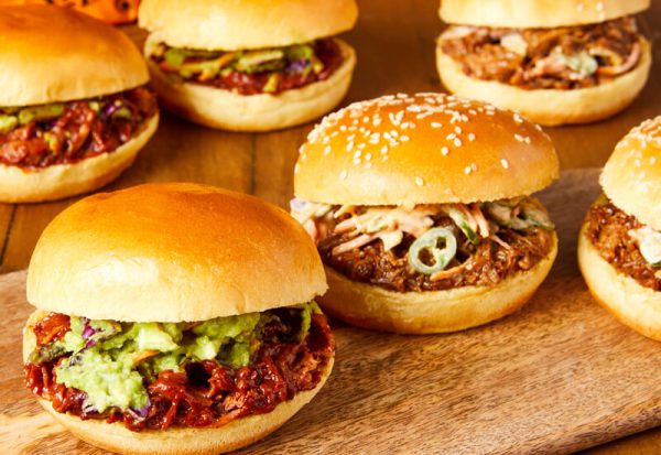 A close up of several burger buns filled with meat and toppings on a wooden serving board