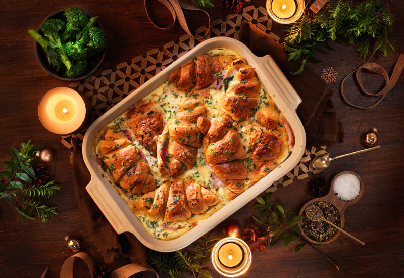 An overhead photo of a croissant savoury bake in a big serving dish surrounded by candles and Christmas decorations