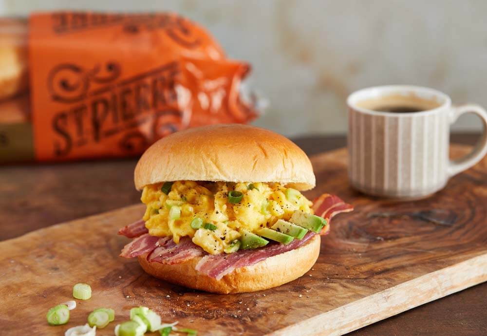 A photo of a burger bun filled with fried bacon, sliced avocado and scrambled egg on a board with a small coffee