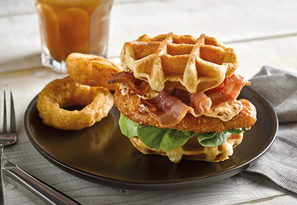 A stack of St Pierre brioche waffles on a plate, with fried bacon, lettuce and fried chicken in between them