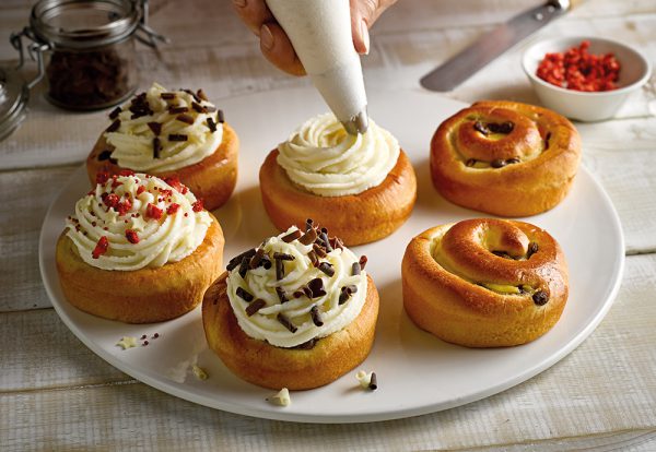 Six St Pierre Brioche Swirls on a plate with a vanilla cream frosting being piped onto one of them