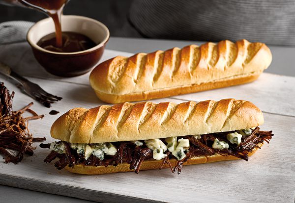 Two St Pierre Soft Brioche Baguettes on a wooden board, with one filled with beef brisket and melted cheese