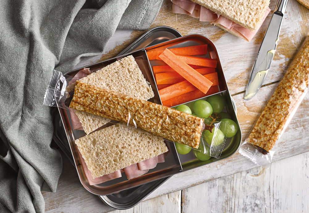 An overhead photo of a lunchbox with sandwiches, crapes and carrot sticks inside, and wrapped chocolate filled crepes on top