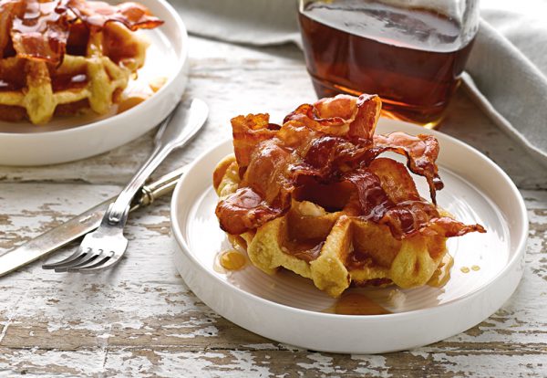 A brioche waffle on a plate with fried bacon and maple syrup on top