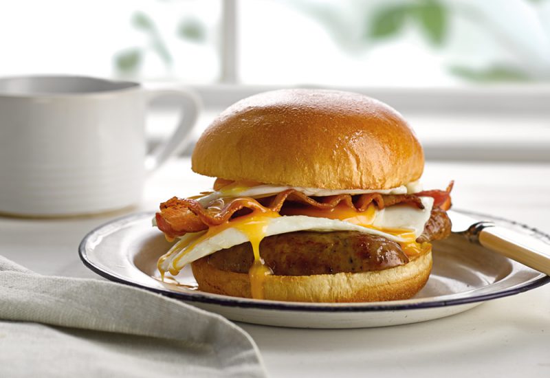 sp_web__0041_breakfast_brioche_burger A close up of a St Pierre Brioche Burger Bun filled with sausages, fried eggs and crispy bacon on a plate with a knife on the side
