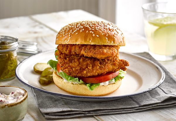 A sesame seeded brioche burger bun filled with fried chicken fillets and salad, served on a plate