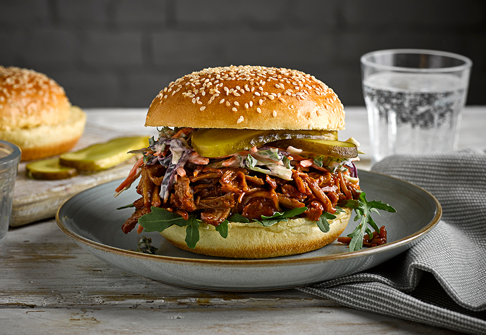 A sesame seeded brioche burger bun filled with pulled pork and salad on a plate on a wooden table