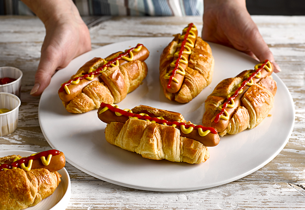 A plate of four St Pierre Brioche Hot Dog Rolls filled with sausages and topped with mustard and ketchup
