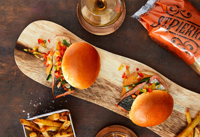 An overhead photo of two burgers on a wooden board with chips, a glass of wine and burger bun pack around the sides