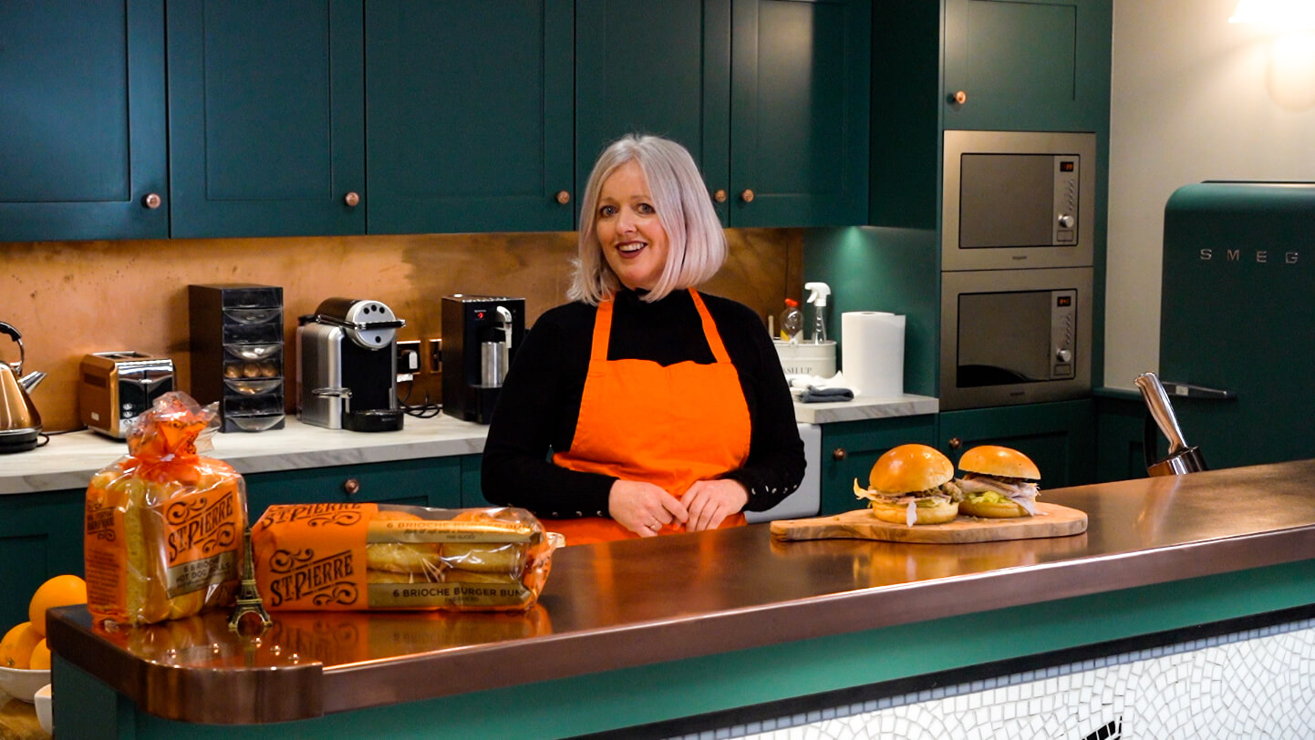A lady in a kitchen wearing an orange apron with St Pierre products and burgers on the countertop